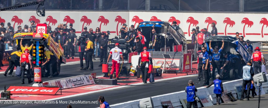 Two drag racing teams prepare their funny cars on the starting line, with crew members working and spectators watching at a racetrack.