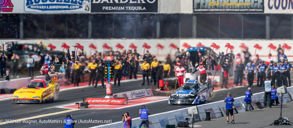 Two drag racing cars on a racetrack prepare to start, with crew members, officials, and spectators standing along the sidelines.