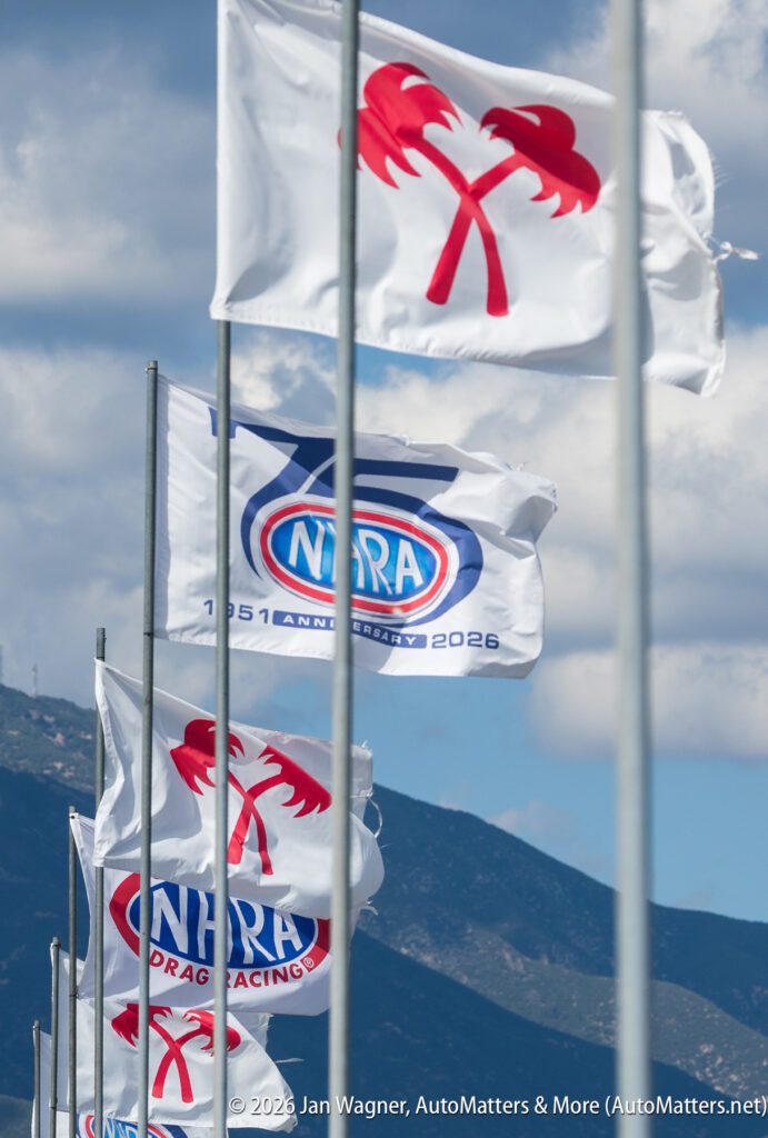 White flags with red palm tree logos and NHRA Drag Racing logos are displayed on flagpoles against a backdrop of mountains and a partly cloudy sky.