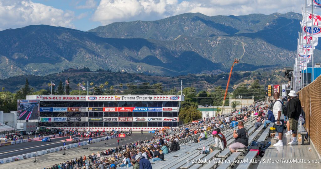 Spectators watch a drag race at a stadium with mountains in the background. The stands are partially filled, and sponsor banners are visible above the track.