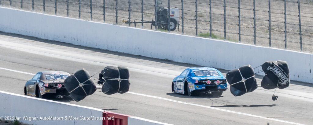 Two race cars deploy parachutes as they slow down on a racetrack after a run, with safety barriers and fencing visible in the background.