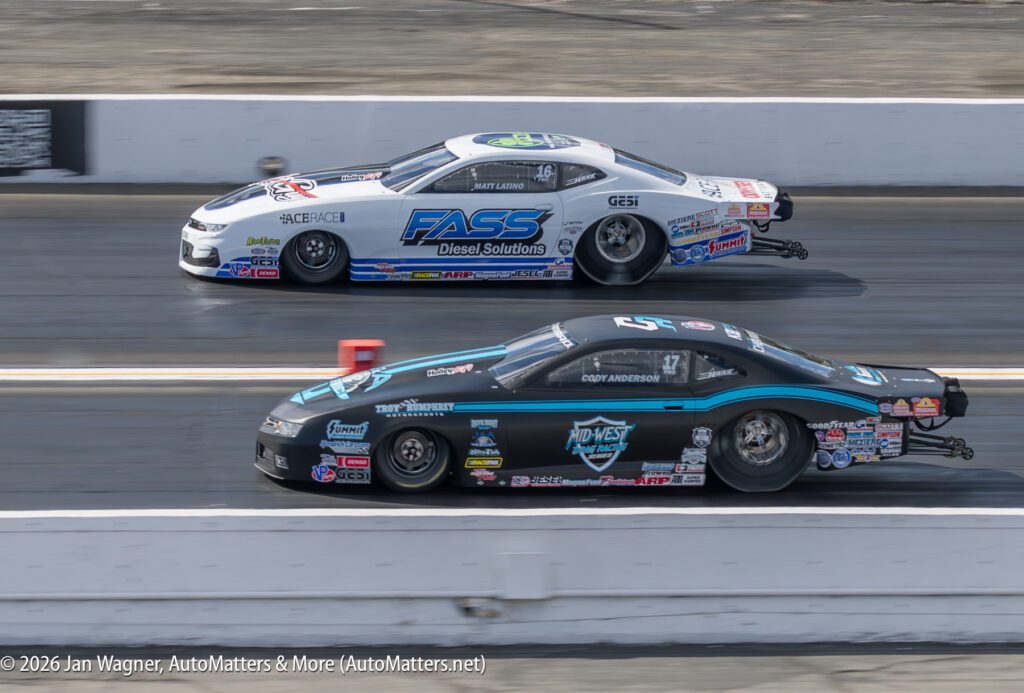 Two drag race cars, decorated with sponsor logos, race side by side on a straight track.