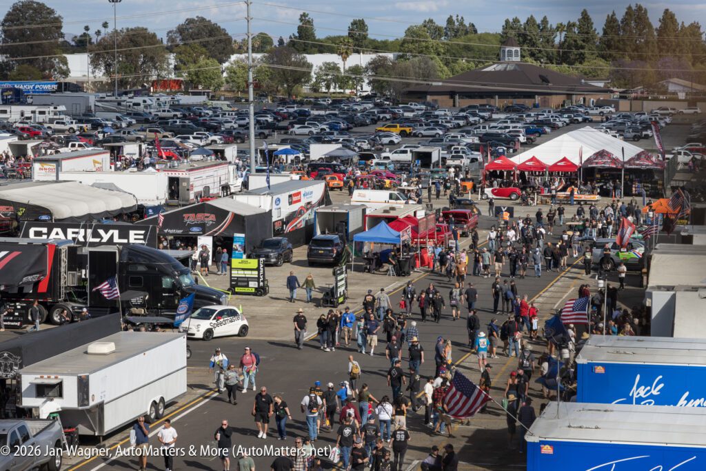 A large outdoor car show with many people walking among various vendor booths, tents, and parked vehicles in a busy parking lot.