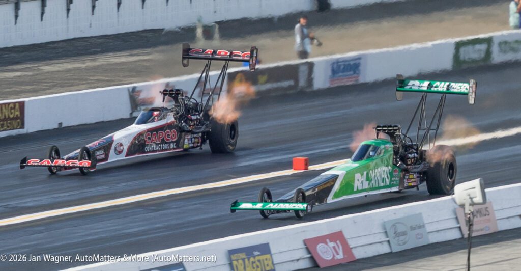 Two dragster race cars accelerate down a dragstrip, with flames visible from their exhausts, as they compete side by side.