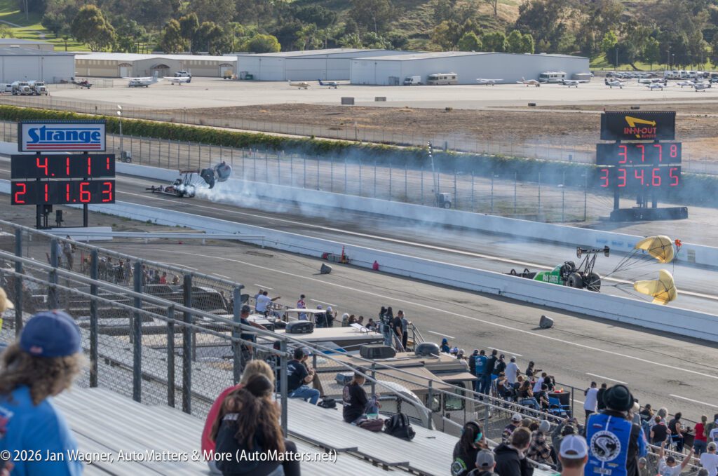 Two drag racing cars speed down a track, one deploying parachutes; electronic displays show elapsed times and speeds as spectators watch from the stands.