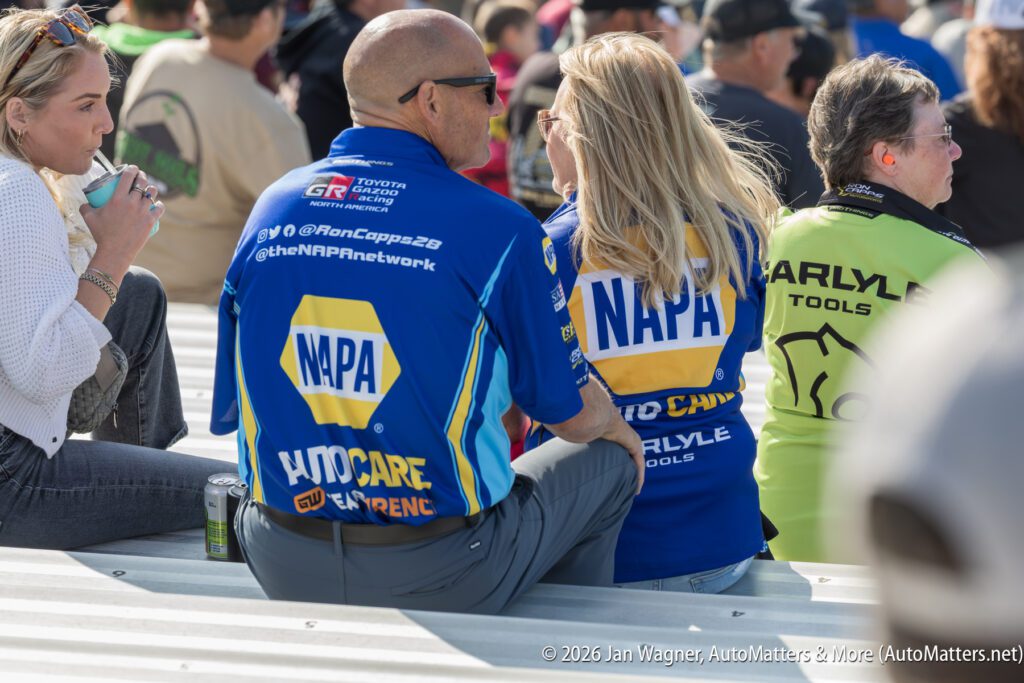 A group of people sit on bleachers at an outdoor event, with two individuals wearing matching blue NAPA AutoCare shirts.