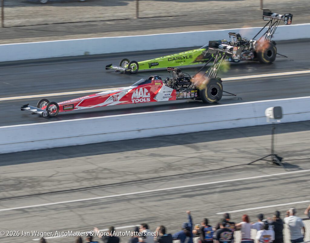 Two top fuel dragsters race side by side on a drag strip, emitting flames from their exhausts as spectators watch from behind the barrier.