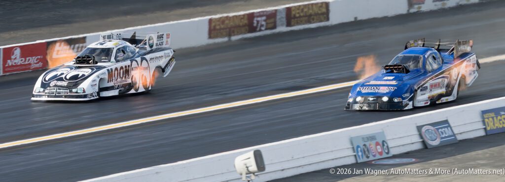 Two funny cars race side by side on a drag strip, with flames visible from the exhausts and sponsor logos on the vehicles.
