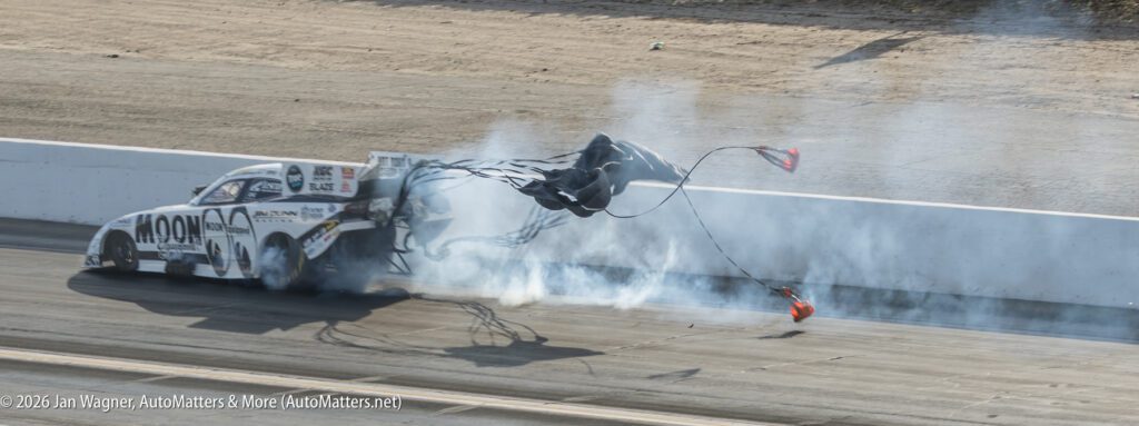 A drag racing car deploys parachutes while smoke trails behind it on a racetrack.