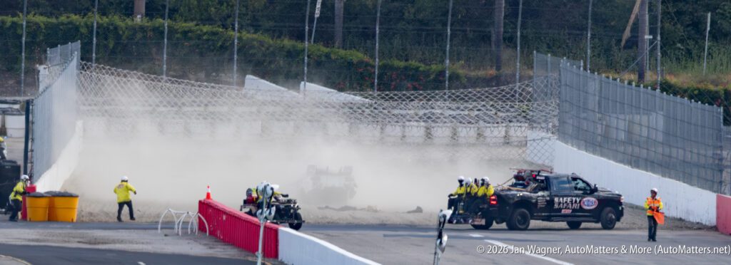 A flipped race car sits in a cloud of dust on a racetrack as safety crews and vehicles respond to the accident.