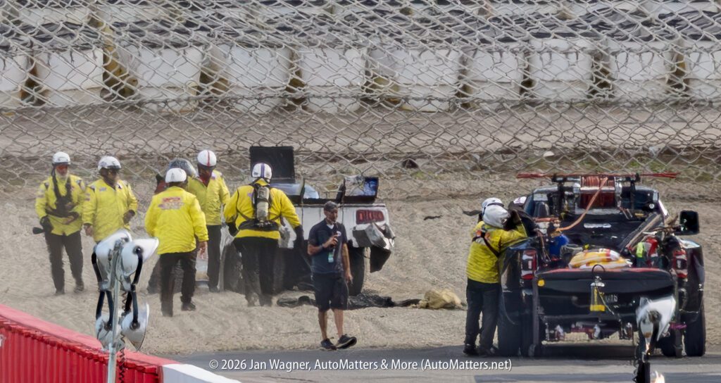 Track safety crew and vehicles attend to an incident on a racetrack, with personnel and equipment gathered near a damaged car behind a protective fence.