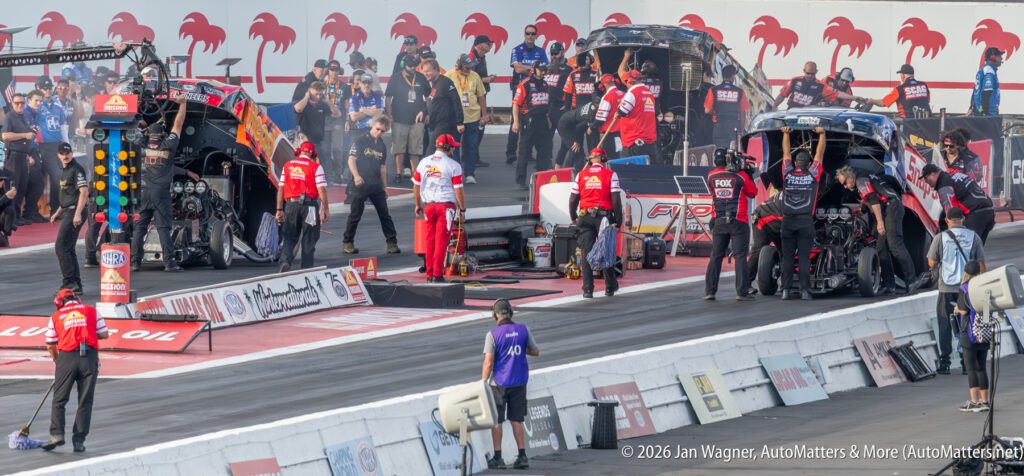 Crew members prepare two drag racing cars at the starting line, with team personnel and equipment visible on both sides of the track.