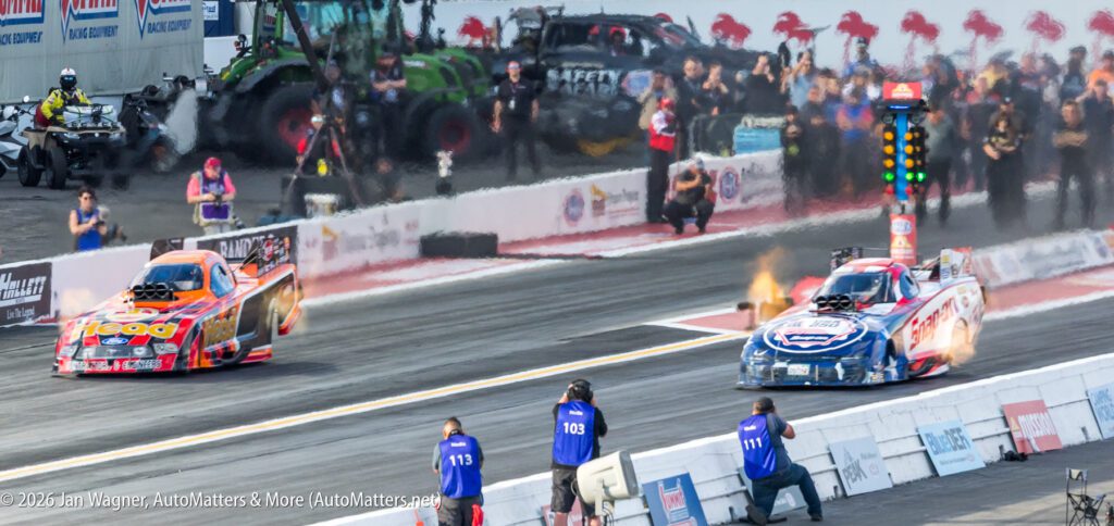 Two drag racing cars accelerate on a track with flames visible from their engines; spectators and crew watch in the background.