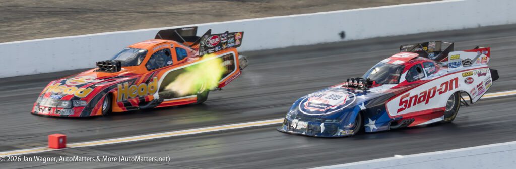 Two drag racing cars speed down a track, with flames visible from the exhaust of the orange car on the left; both cars display sponsor logos.