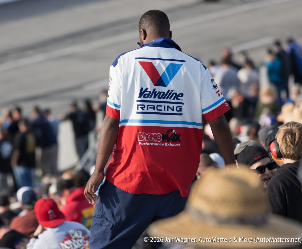 A man in a Valvoline Racing shirt stands among a crowd at an outdoor event, with a racetrack visible in the background.