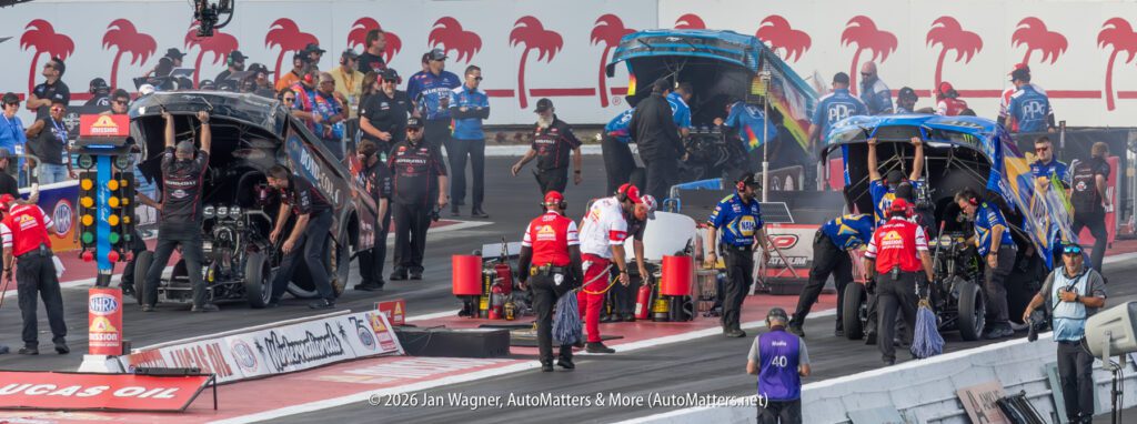 Two drag racing cars with raised bodies are serviced by pit crews on a racetrack, with race officials and crew members standing nearby. Palm tree silhouettes decorate the background wall.