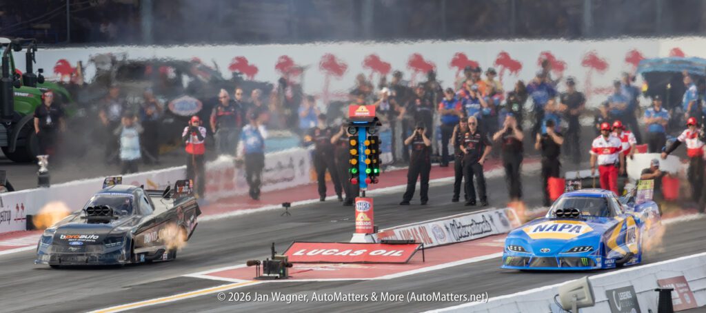 Two drag racing cars accelerate from the starting line, flames visible from exhausts, with crew members and spectators watching in the background.