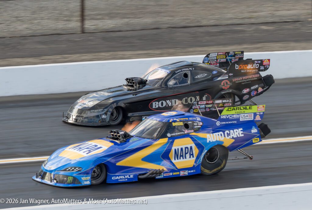 Two funny cars, one in blue and yellow NAPA livery and one in black Bond-Coat livery, race side by side on a drag strip, both emitting flames from their exhaust.