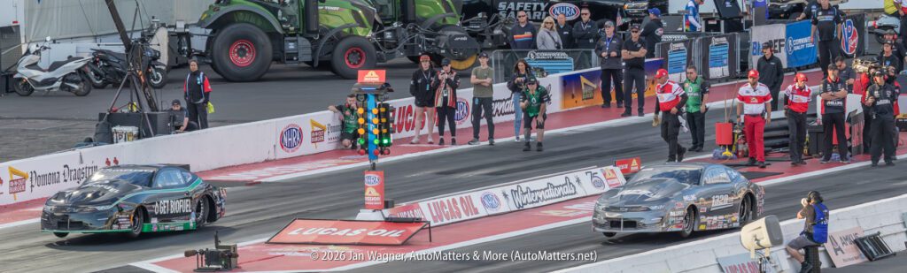 Two drag racing cars at the starting line on a racetrack, surrounded by officials, crew members, and safety personnel, with equipment and vehicles in the background.