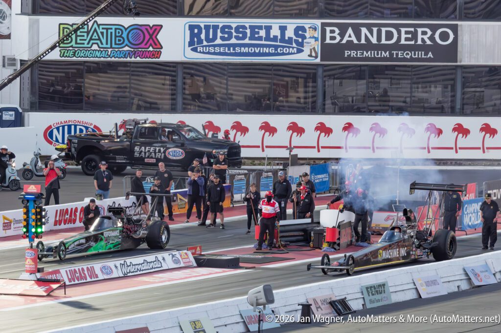 Two dragsters line up on a racetrack with crew members and officials nearby, as safety vehicles and advertisements are visible in the background.