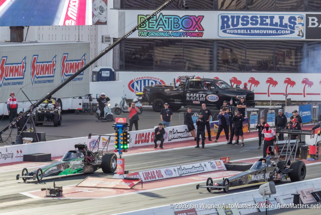 Two drag racing cars at the starting line on a racetrack, with crew members, officials, and a safety truck in the background.