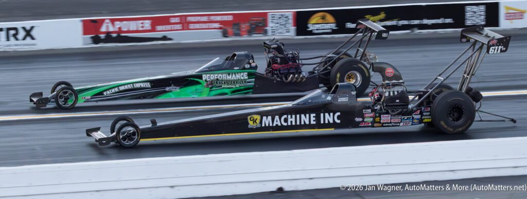 Two drag racing cars speed side by side down a racetrack, with sponsors' logos visible on the vehicles and track barriers in the background.
