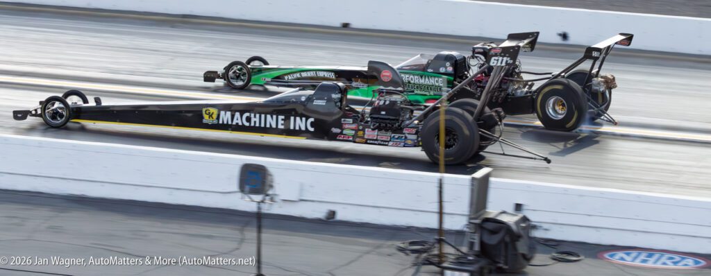 Two dragster race cars accelerate side by side down a drag strip during a daytime NHRA event.