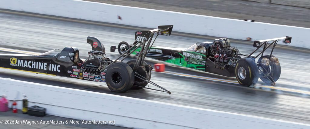 Two dragster cars racing side by side on a track, with smoke visible near the rear tires.