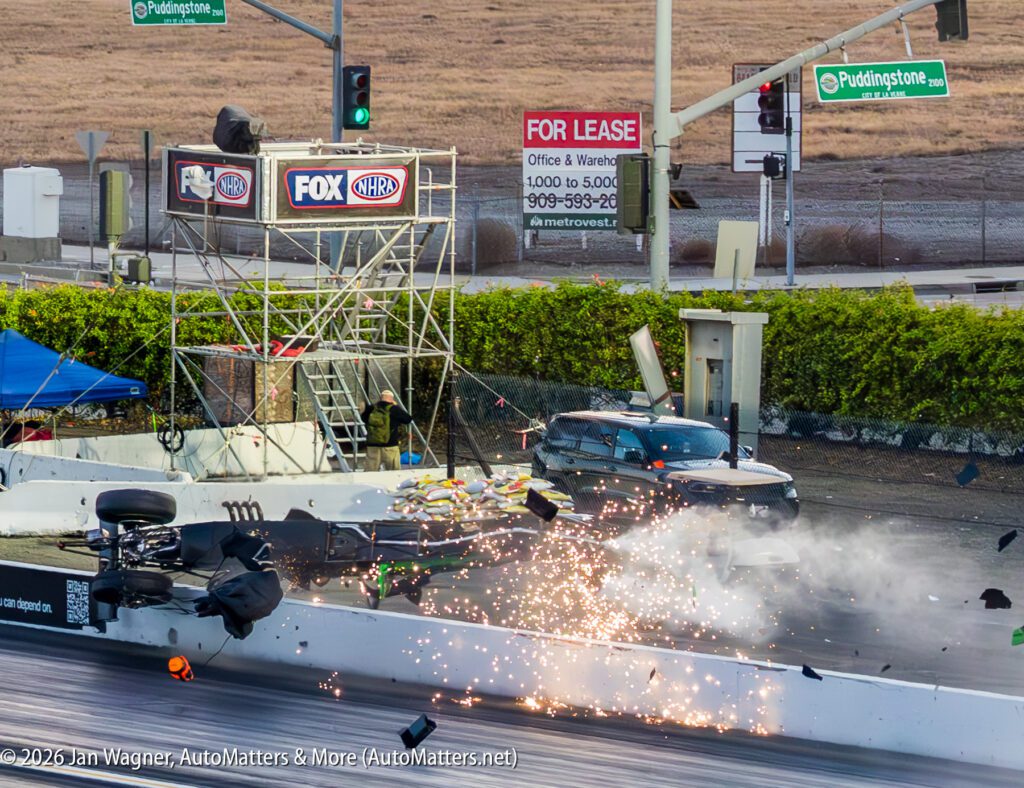 A race car crashes near the track barrier, debris and sparks flying, with a black vehicle and camera equipment in the background near "For Lease" signs and streetlights.