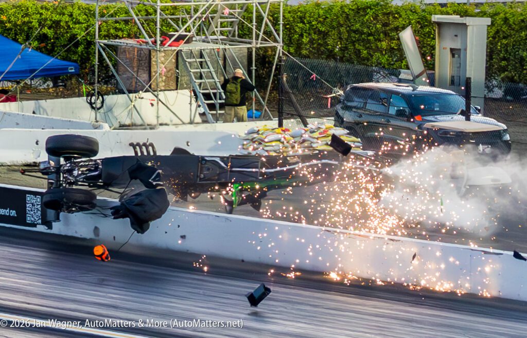 A race car crashes into a barrier at high speed, sending debris and sparks into the air as emergency personnel observe from behind a fence.