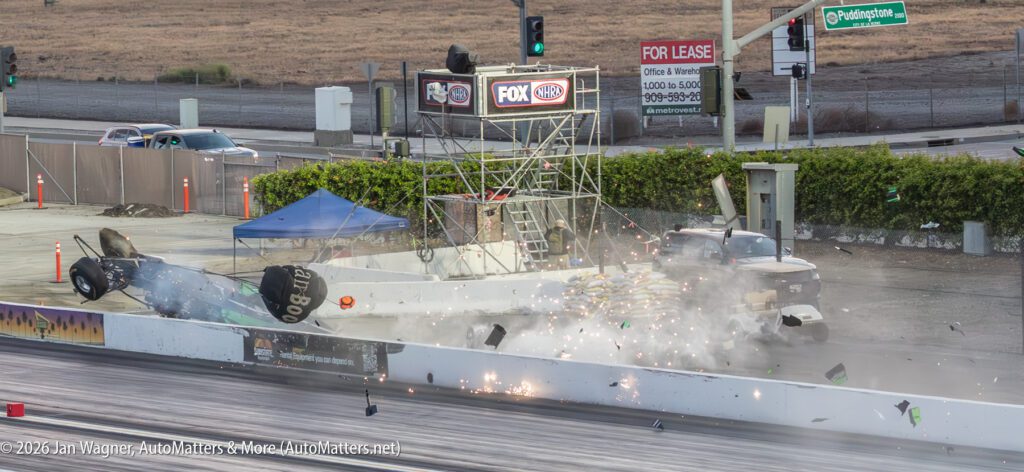 A race car crashes and flips over a concrete barrier at a drag strip, debris flying as bystanders watch from a tower; a pickup truck is struck by the car.
