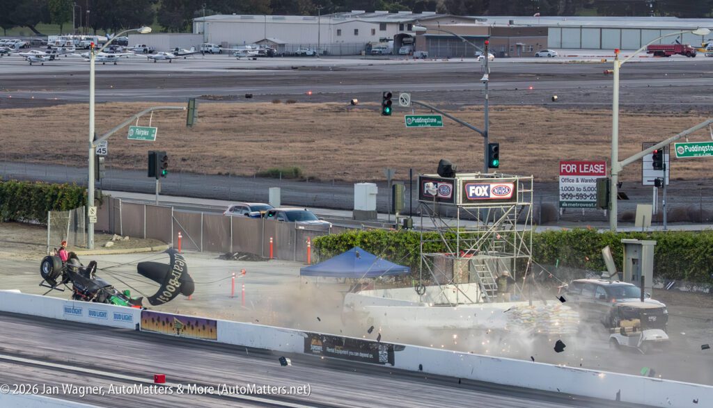 A drag racing car crashes and flips upside down near the finish line, debris and dust in the air, while emergency personnel respond at a racetrack.