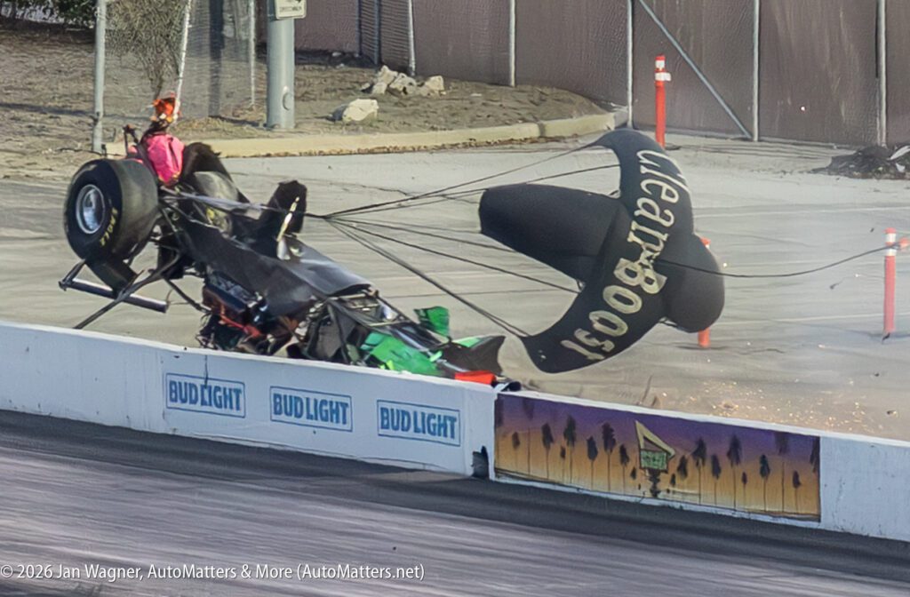 A drag racing car crashes into a barrier, its front end raised and rear parachute deployed, with debris and smoke visible around the impact site.