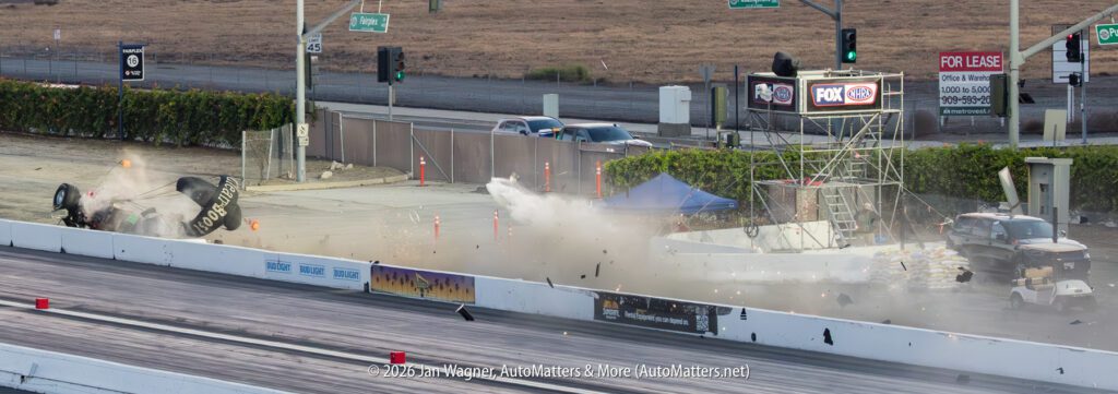 A drag racing car crashes and collides with a camera stand near the track, sending debris and dust into the air.
