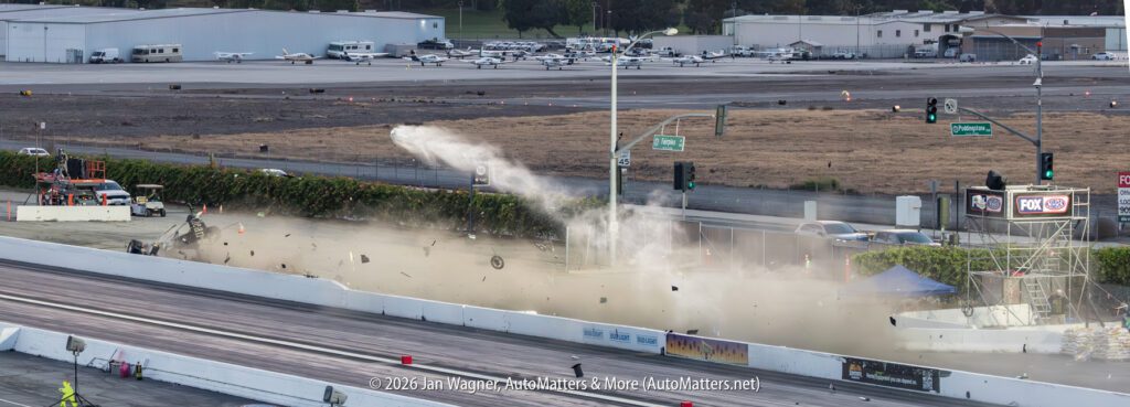 A race car crashes through a barrier at a drag strip, sending debris and dust into the air; a nearby airport and runway are visible in the background.