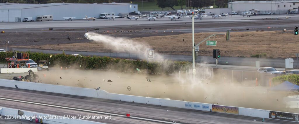 A race car crash at a drag strip sends debris and dust into the air, with scattered parts visible on the track and spectators in the background.