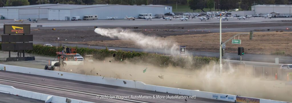 A jet-powered dragster crashes at a racetrack, sending debris and smoke flying across the track while spectators and vehicles appear in the background.