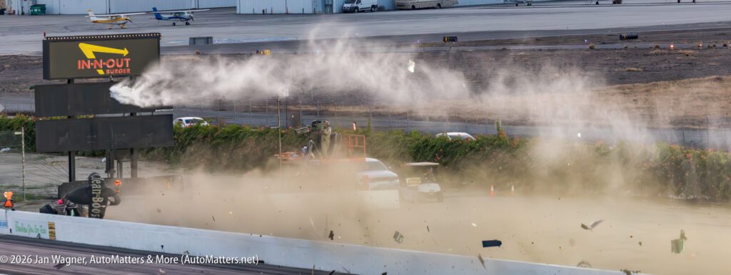 A drag racing car crash sends debris, dust, and smoke into the air near an In-N-Out Burger track sign at an airport drag strip.