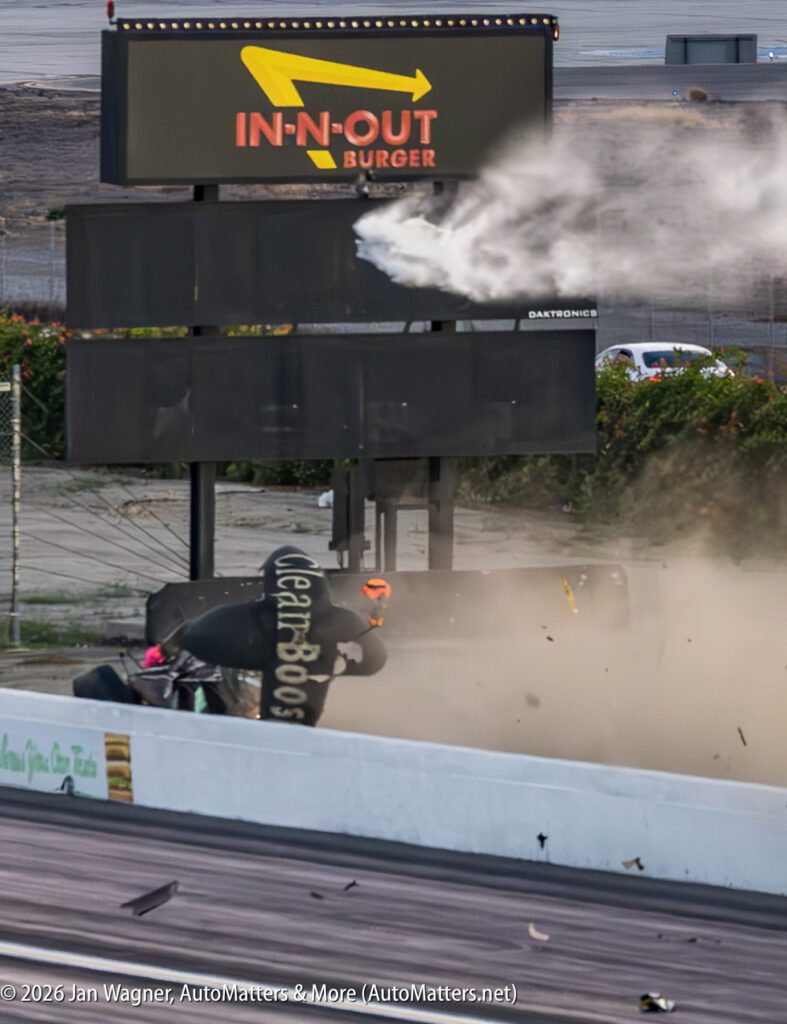 A dragster crashes and flips upside down at a racetrack, with debris and dust airborne beneath an In-N-Out Burger sign.