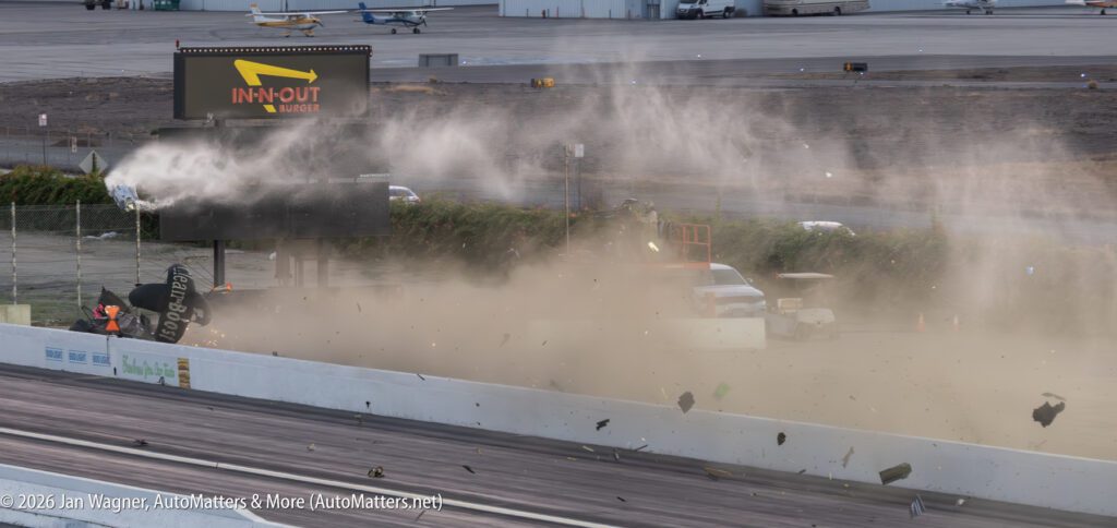 A drag racing crash sends debris and dust into the air near the track, with a damaged sign and scattered objects visible.