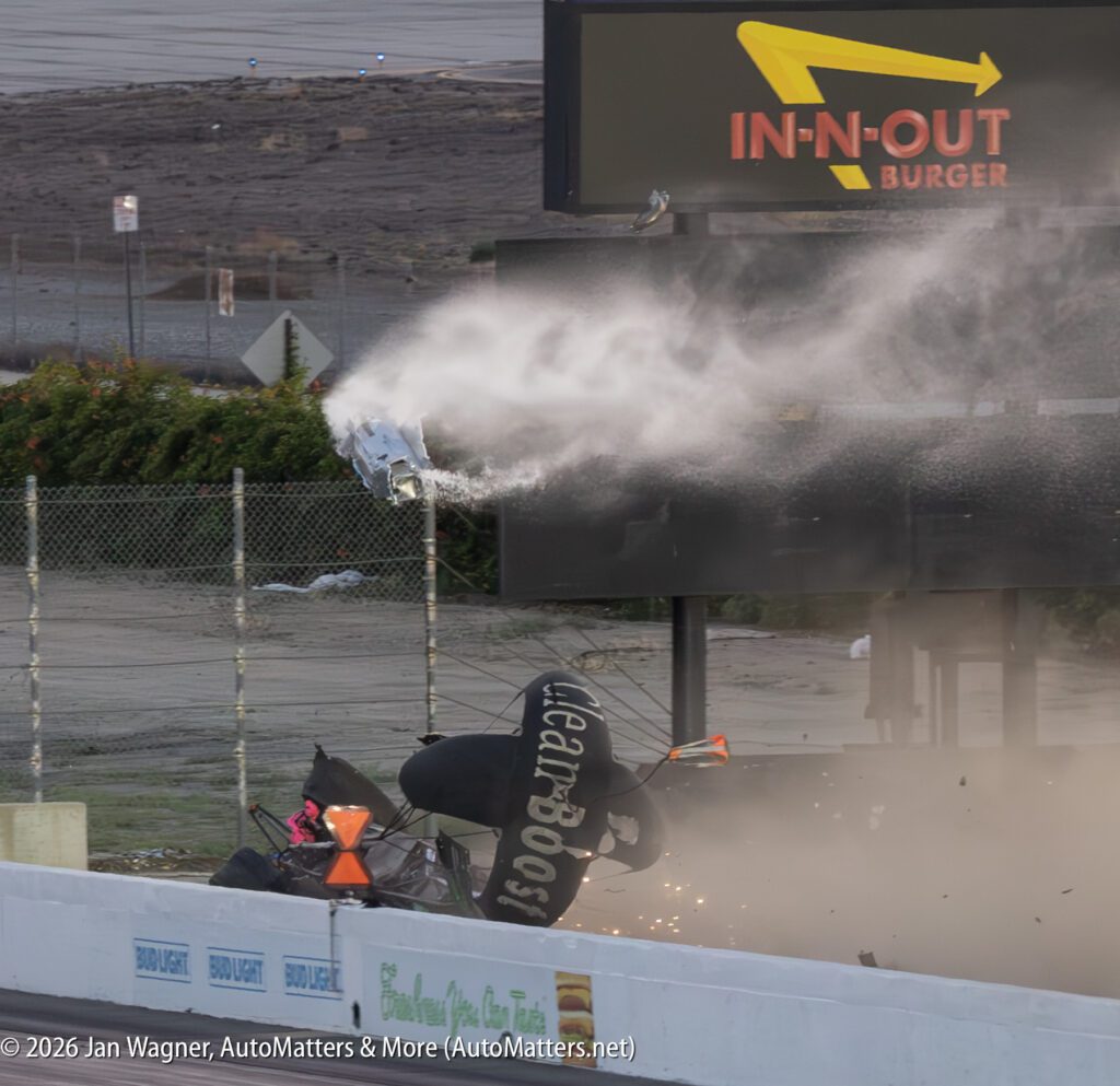 A race car crashes into a barrier, sending debris and smoke into the air near an In-N-Out Burger sign at a racetrack.