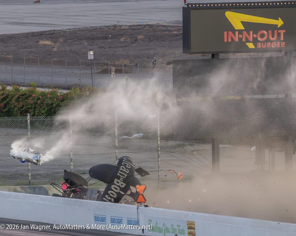 A race car crashes into a barrier at a racetrack, creating debris and a cloud of dust, with an In-N-Out Burger sign visible in the background.