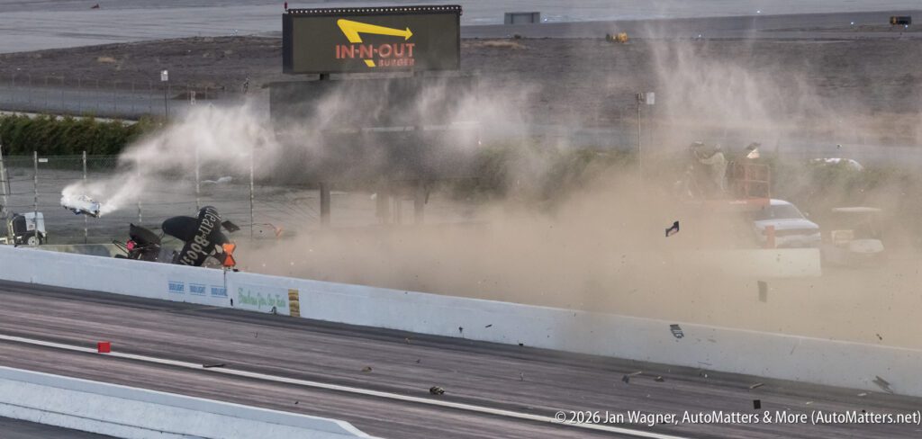 A drag racing car crashes, sending debris and dust into the air near a racetrack barrier; an IN-N-OUT Burger sign is visible in the background.