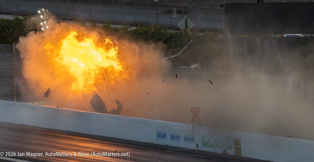 A fiery explosion erupts on a racetrack, sending debris and smoke into the air near a barrier wall.