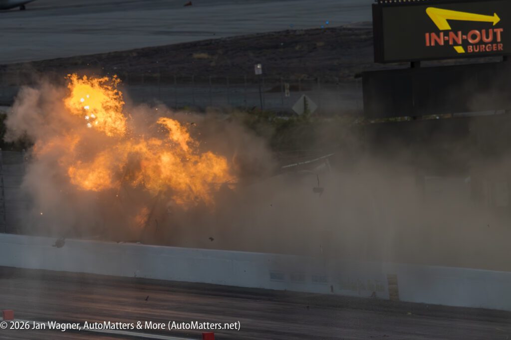 A large fireball and cloud of smoke erupt from behind a barrier on a racetrack, with an In-N-Out Burger sign visible in the background.