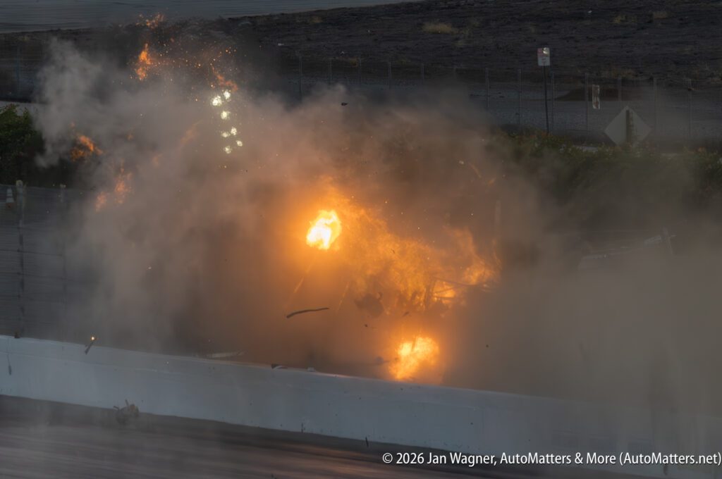 A fiery car crash with flames and debris filling the air on a racetrack, partially obscured by smoke and fire.