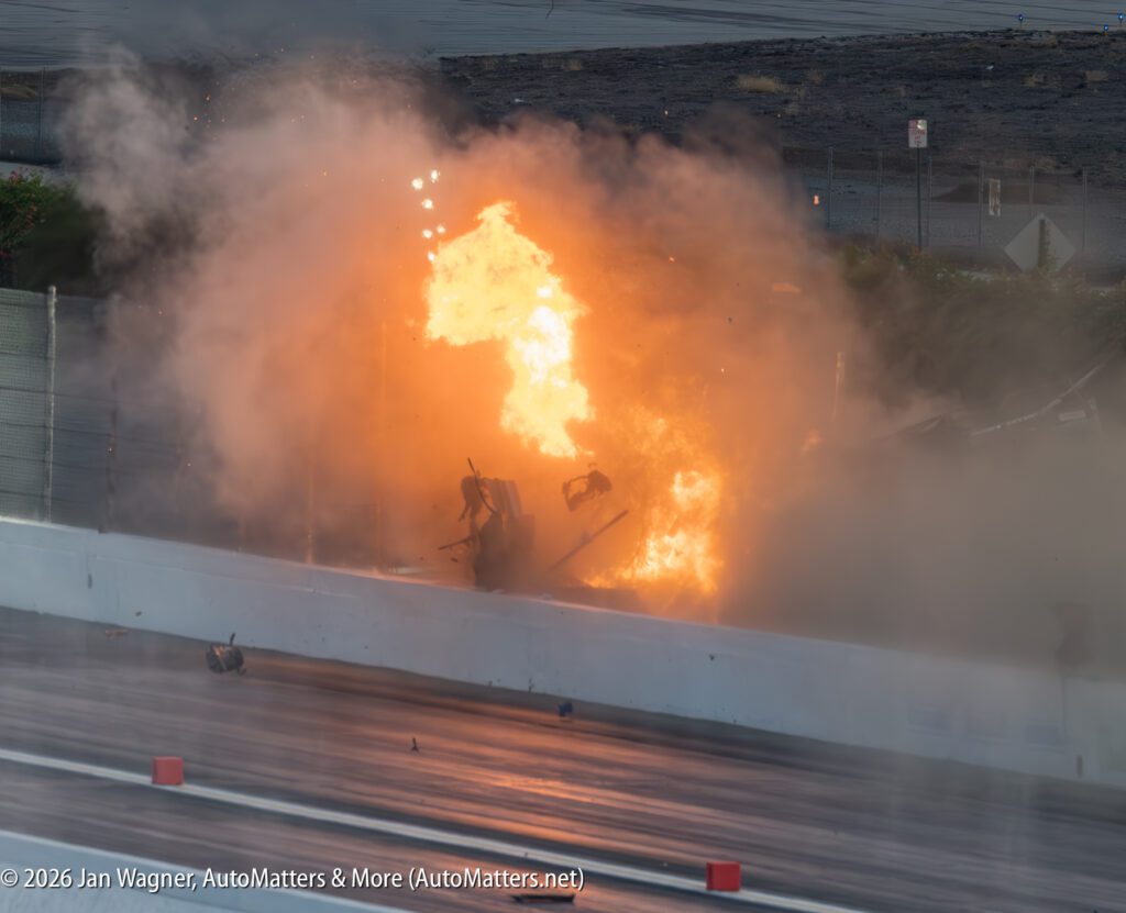 A race car collides with a barrier and bursts into flames on a drag strip, with debris and smoke filling the air.