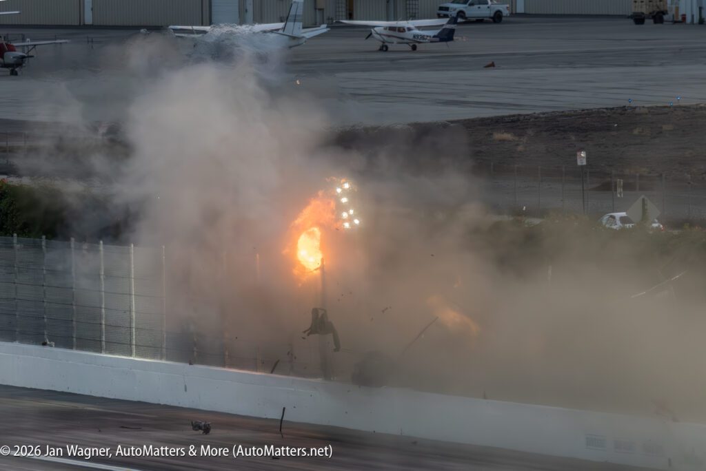 A burst of flames, smoke, and debris rises from a crash against a racetrack barrier, with airplanes and hangars visible in the background.