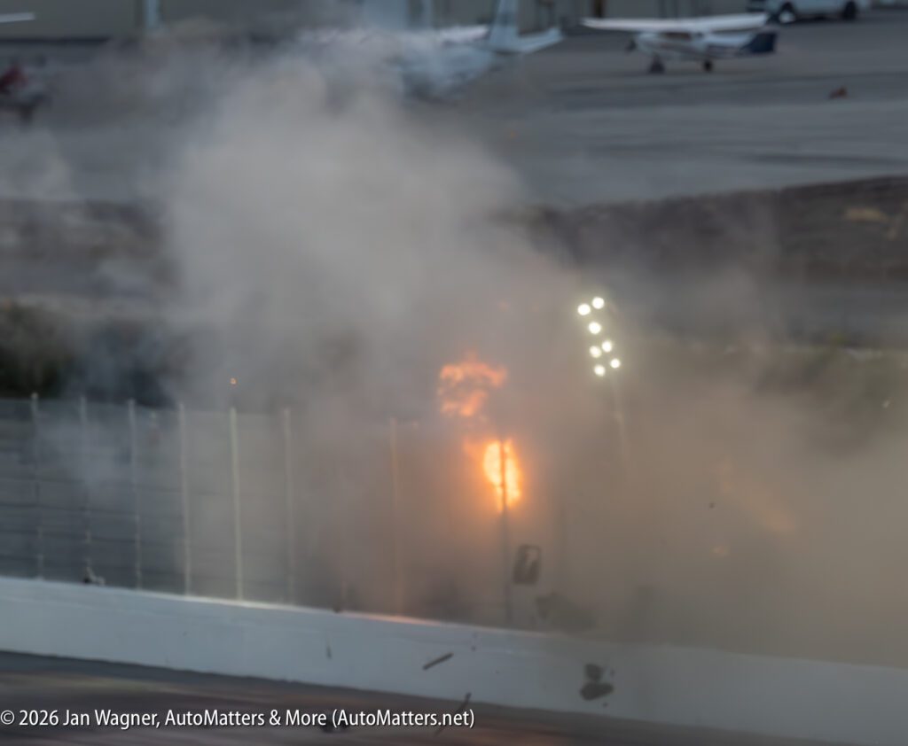 A crash with visible flames and smoke against a barrier on a racetrack, with blurred airport area and planes in the background.