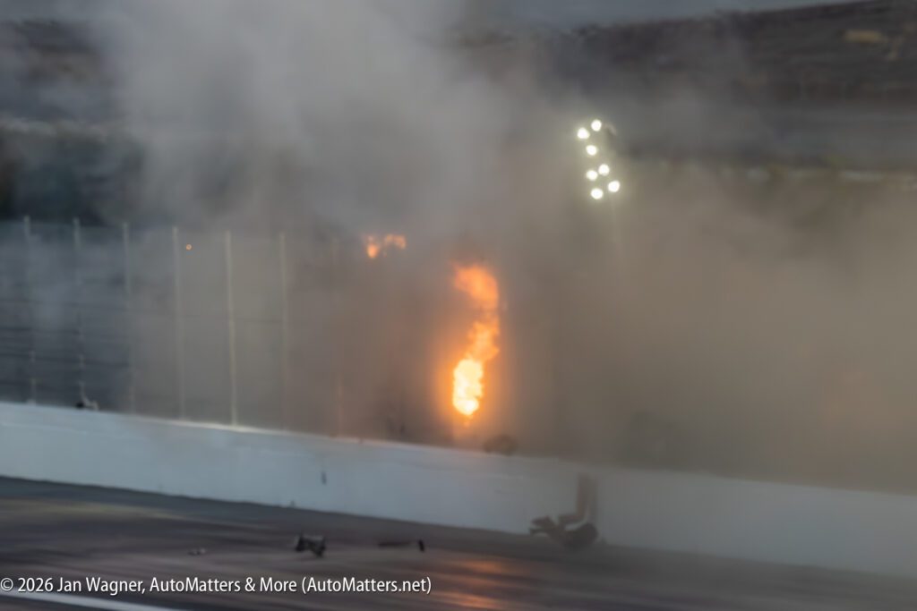 A fire burns behind a racetrack barrier with thick smoke rising; debris and a helmet are visible on the track in the foreground.